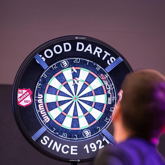 Ein Spieler wirft einen Pfeil auf ein beleuchtetes Dartboard mit dem Schriftzug „GOOD DARTS“ im Hintergrund.